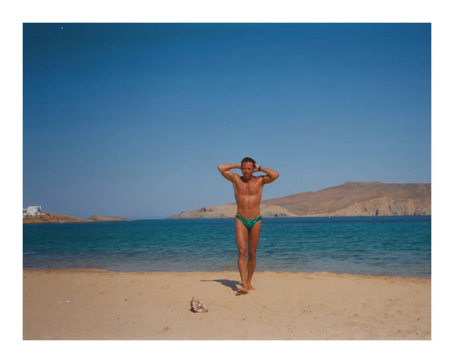 A muscular, tanned man posing center-frame on a sandy beach. He is wearing green Speedo-style swim briefs, standing barefoot with both hands behind his head. The background shows blue ocean water and large, arid mountains under a clear sky.