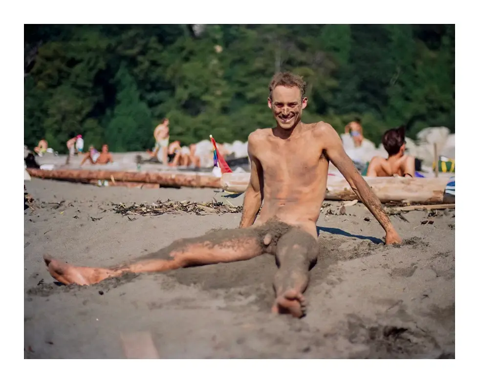 A shirtless, bearded man sitting nude and smiling broadly on a sandy beach. He is leaning back, supported by his hands on the dark sand, with visible dirt on his body. The background shows a large log, other blurred beachgoers, and green foliage under natural light.