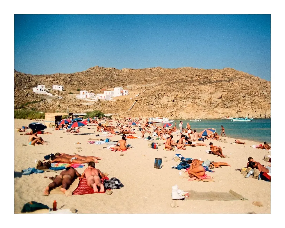 Crowded vintage photograph of a Mykonos, Greece beach. Dozens of nude and semi-nude men sunbathe on the sand. White-washed Cycladic houses dot the rocky hill in the background under a clear blue sky.