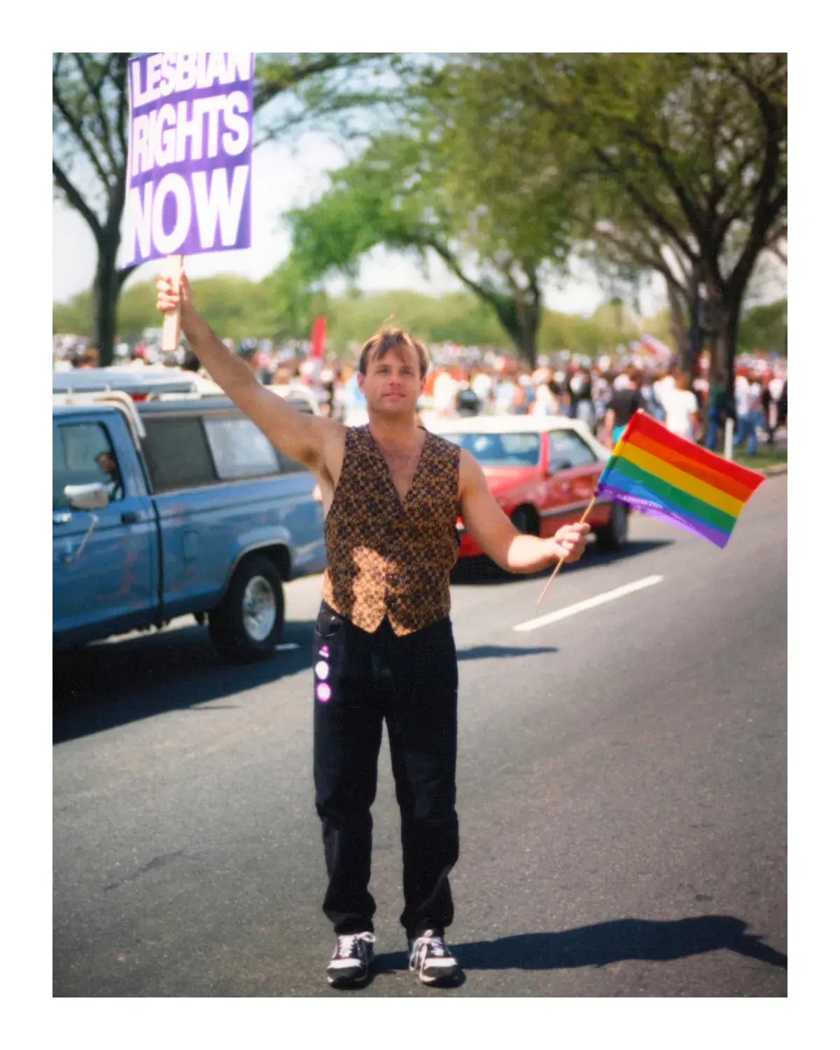 A gay rights march shows a man standing in a street. He wears a patterned vest and dark pants, holding a sign in one raised hand reading "LESBIAN RIGHTS NOW" and a small rainbow flag in his other hand.