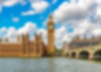 Big Ben and the Houses of Parliament under a blue sky with clouds, next to the River Thames and Westminster Bridge. Bright and vibrant scene.