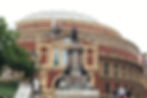 A red brick building with ornate details and a dome in the background. A statue in the foreground. Three people stand nearby. Cloudy sky.