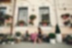 Woman and girl in black dresses sit at a pink table outside a quaint cafe. Surrounded by potted plants and colorful decor, enjoying a chat.