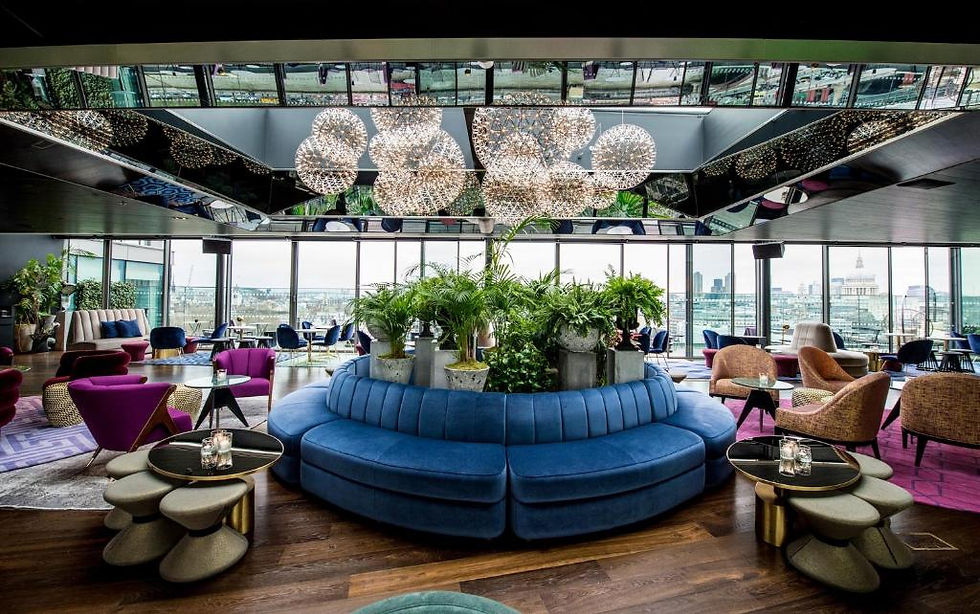 Luxurious lounge with blue circular seating, plants, modern chandeliers, and panoramic city views. Vibrant chairs add colorful accents.