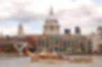 Ornate boat with flags on the Thames, rowed by people. Background features St. Paul's Cathedral and modern buildings under cloudy sky.