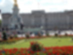 People walk in front of Buckingham Palace, with colorful flower gardens and the Victoria Memorial statue. Bright and sunny atmosphere.