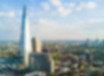 Aerial view of the Shard in London under a partly cloudy sky. The modern glass skyscraper towers over surrounding buildings and cityscape.
