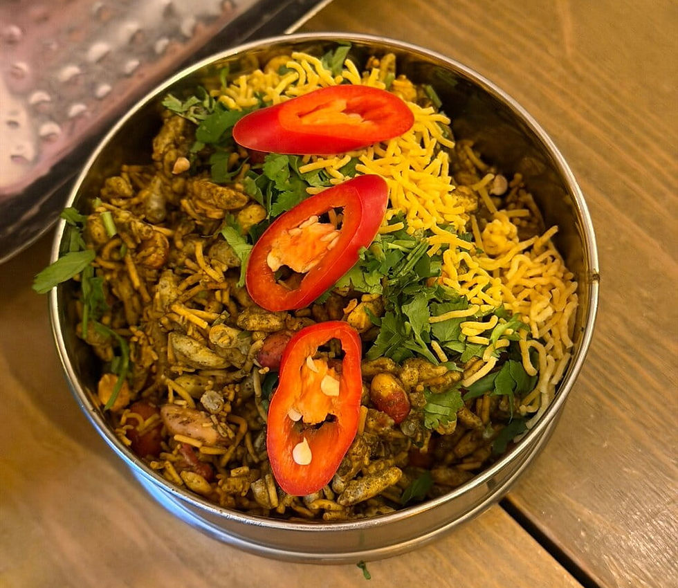 Bowl of Indian bhel puri topped with sliced red peppers, sev, and cilantro on a wooden table. Vibrant colors and fresh ingredients.