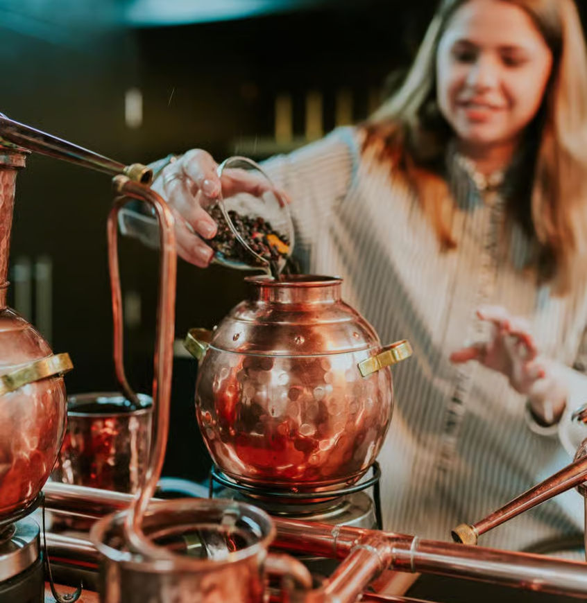A person pours ingredients into a copper pot in a workshop, during the gin-making class with tasting in the Piston Distillery, Cheltenham surrounded by distillation equipment. Soft lighting creates a warm atmosphere.