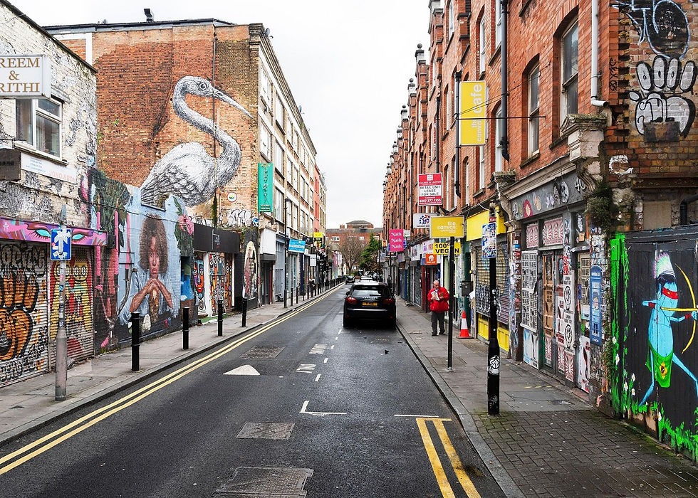 Street with vibrant murals, including a large bird. Brick buildings line the road. A person in red walks, and parked cars are visible.