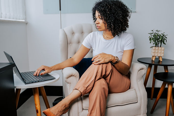 Mulher negra com cabelos crespos cacheados até os ombros, vestindo uma blusa branca e calça marrom, sentada com as pernas cruzadas em uma poltrona, está com o seu braço direito estendido para o notebook e estáolhando para a tela do notebook que está ao seu lado apoiado em uma mesa de canto.