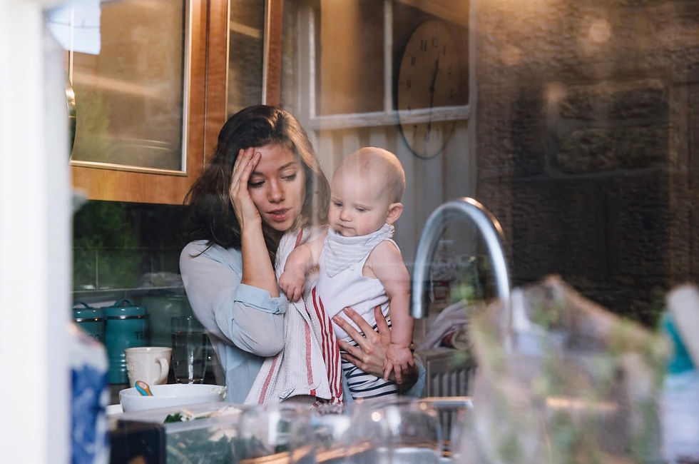 A tired woman holds a baby in a cluttered kitchen, reflecting stress. Wooden cabinets, a clock, and kitchenware are visible in the background.