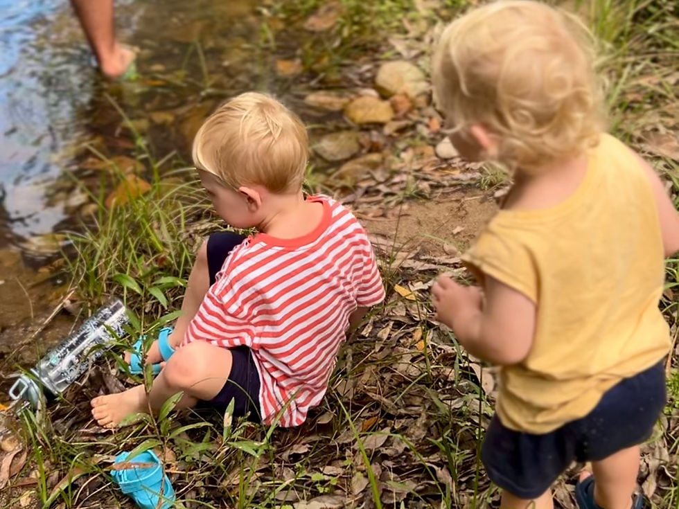 Two children by a stream, one in a striped shirt sitting with a bottle, the other standing. Greenery and water create a playful mood.