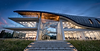 Architectural dusk photograph of the modern STEM building at St Faith's School, Cambridge, showcasing a large curving undulating aerodynamic zinc roof profile, a fully illuminated glass curtain wall facade, and an interior lit helical spiral staircase designed by Cavaleri Partnership.