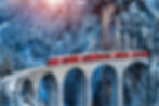 Red Swiss train crossing a stone viaduct through a snowy forest near St. Moritz, with rocky cliffs and frosted trees surrounding the scene.
