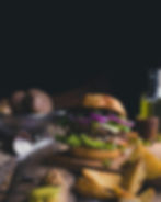 Close-up of a gourmet burger with fresh lettuce, onions, tomato, and cheese, served with rustic potato wedges on a wooden board.