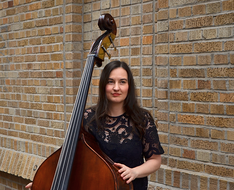 Kate holding an upright bass, standing in front of a brick wall.