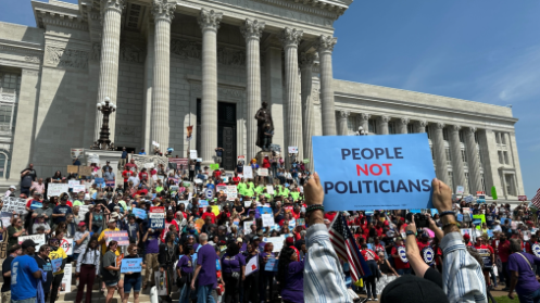 Protesters gather on the steps of the Missouri Capitol, holding signs that demand to prioritize people's needs over political agendas.