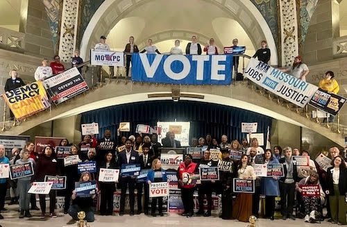 A large group of people gather in the Missouri Capitol Rotunda, holding signs advocating for voting rights and democracy, while prominently displaying a "VOTE" banner. The diverse crowd represents a unified call to action for civic engagement and justice in Missouri.