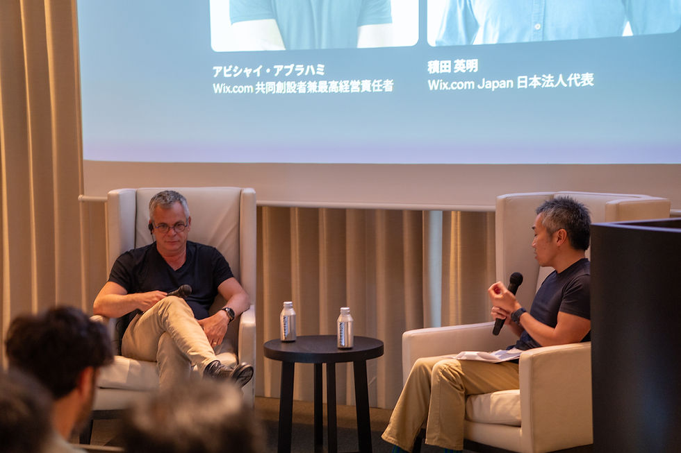 2人が椅子に座り会話中。1人はマイクを持つ。背景にスクリーンがあり、日本語のテキストが表示。リラックスした雰囲気。