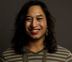 headshot (courtesy of Dena Denny), part of a series of genderqueer and nonbinary folx. i'm wearing a deep red lipstick with shoulder length hair and a gold and silver chain necklace that drops just out of frame. i'm wearing long sleeve t-shirt, gree a horizontally striped shirt, grey-green and brown horizontal stripes