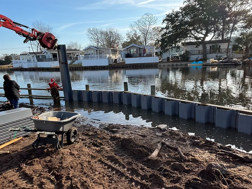 Se están instalando tablestacas de vinilo a lo largo de un canal utilizando una excavadora, guiando un panel en aguas poco profundas y colocando los materiales en la orilla fangosa.