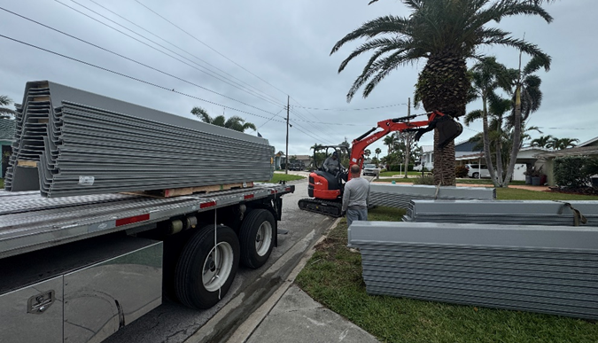 Miniexcavadora descargando tablestacas corrugadas desde un camión de plataforma en una zona residencial tropical.