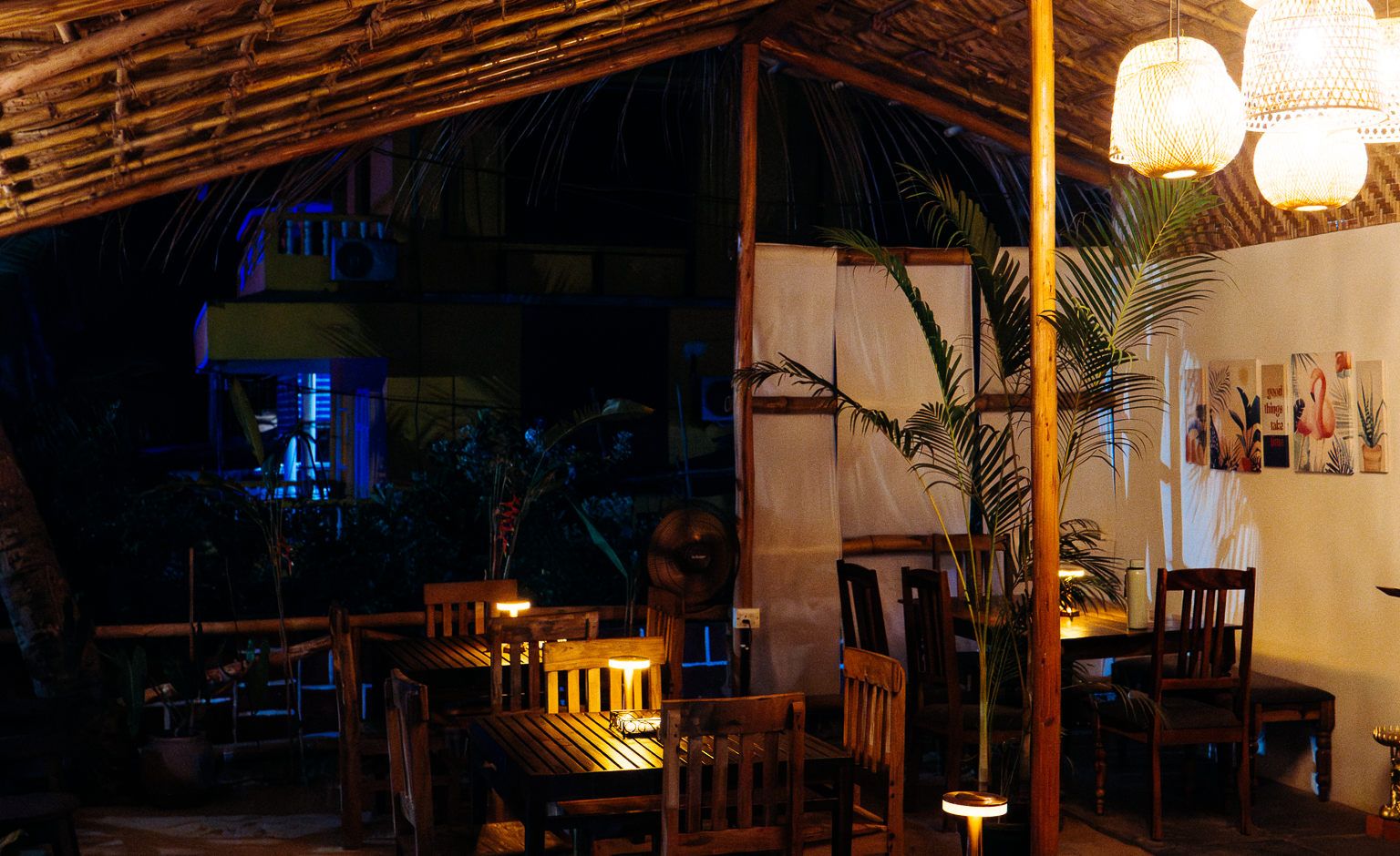 Cozy tropical restaurant with thatched roof, hanging lanterns, wooden tables and chairs on a sand floor, warm glow.
