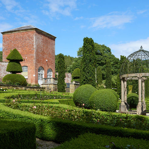 The Walled Garden at Hopton Hall with manicured hedges and topiary around the red brick Summerhouse, under a bright blue sky, with a wrought-iron gazebo
