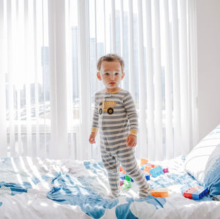 Toddler in striped pajamas stands on a bed with blue sheets, surrounded by colorful toy blocks. Sunlight filters through vertical blinds.