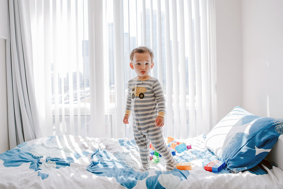 Toddler in striped pajamas stands on a bed with blue sheets, surrounded by colorful toy blocks. Sunlight filters through vertical blinds.