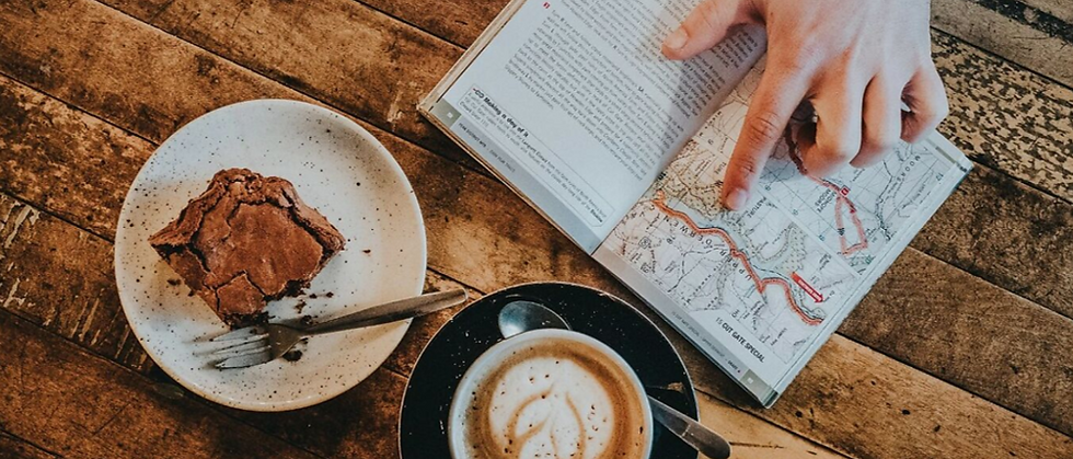 Hand points at a map in a book on a rustic wooden table. Nearby are a cappuccino and a chocolate brownie on ceramic plates. Cozy ambiance.