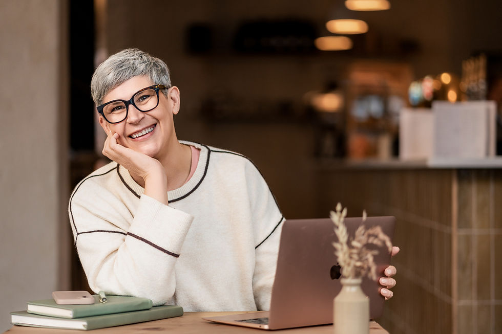 Sally Goodwin, smiling with gray hair and glasses sits in a cozy cafe, hand on cheek, next to a laptop and notebooks. Warm lighting sets a relaxed mood.