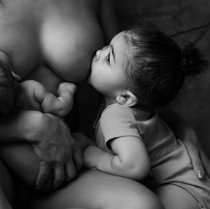 A woman breastfeeds an infant on the left while a toddler nurses from her right breast. The black and white setting is intimate and serene.