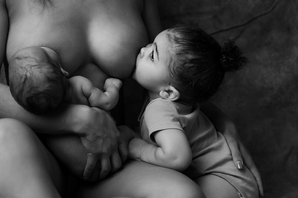 A woman breastfeeds an infant on the left while a toddler nurses from her right breast. The black and white setting is intimate and serene.