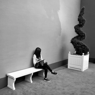 Dans un hall une jeune femme est assise sur un banc blanc, un arbre est posé à coté d'elle dans une boite.