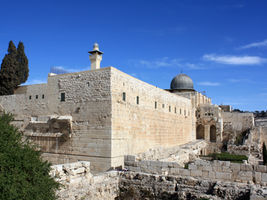 Stone religious complex with a gray dome and minaret under a bright blue sky, viewed from below.