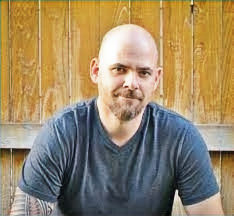 Bald man with a goatee and tattoos on his arm, wearing a blue shirt, smiles slightly against a wooden fence background. Warm, casual mood. Author Cully Perlman
