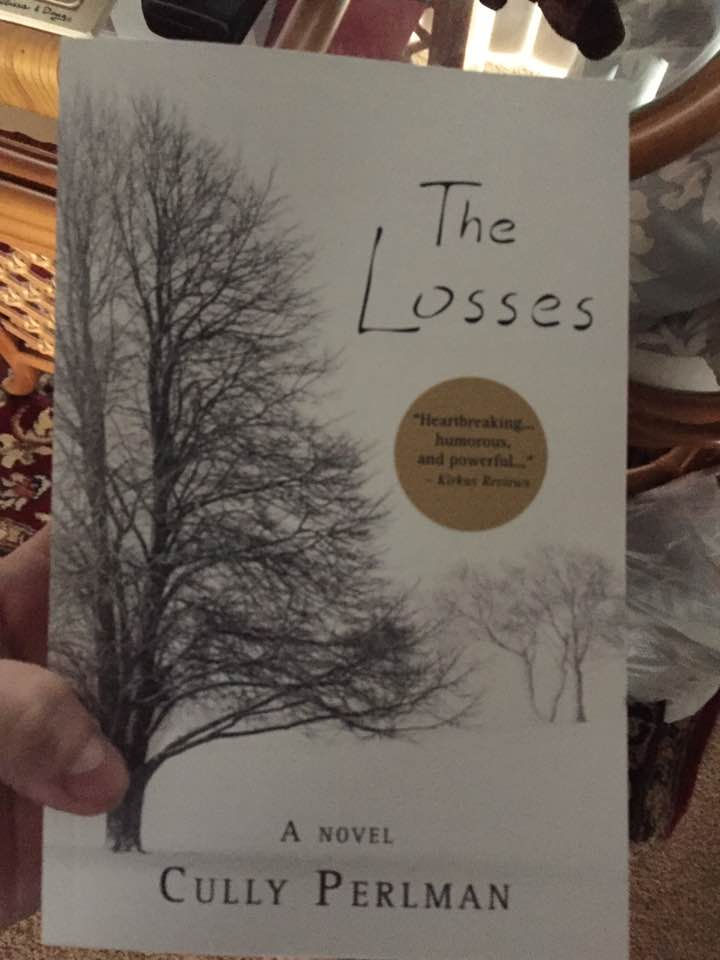 A hand holds "The Losses" novel by Cully Perlman. Cover shows leafless trees. Gold sticker reads, "Heartbreaking, humorous, and powerful."