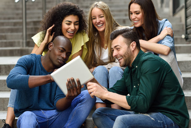 Five friends sit on steps, smiling and looking at a tablet together. They're wearing casual clothes, creating a cheerful, relaxed vibe.