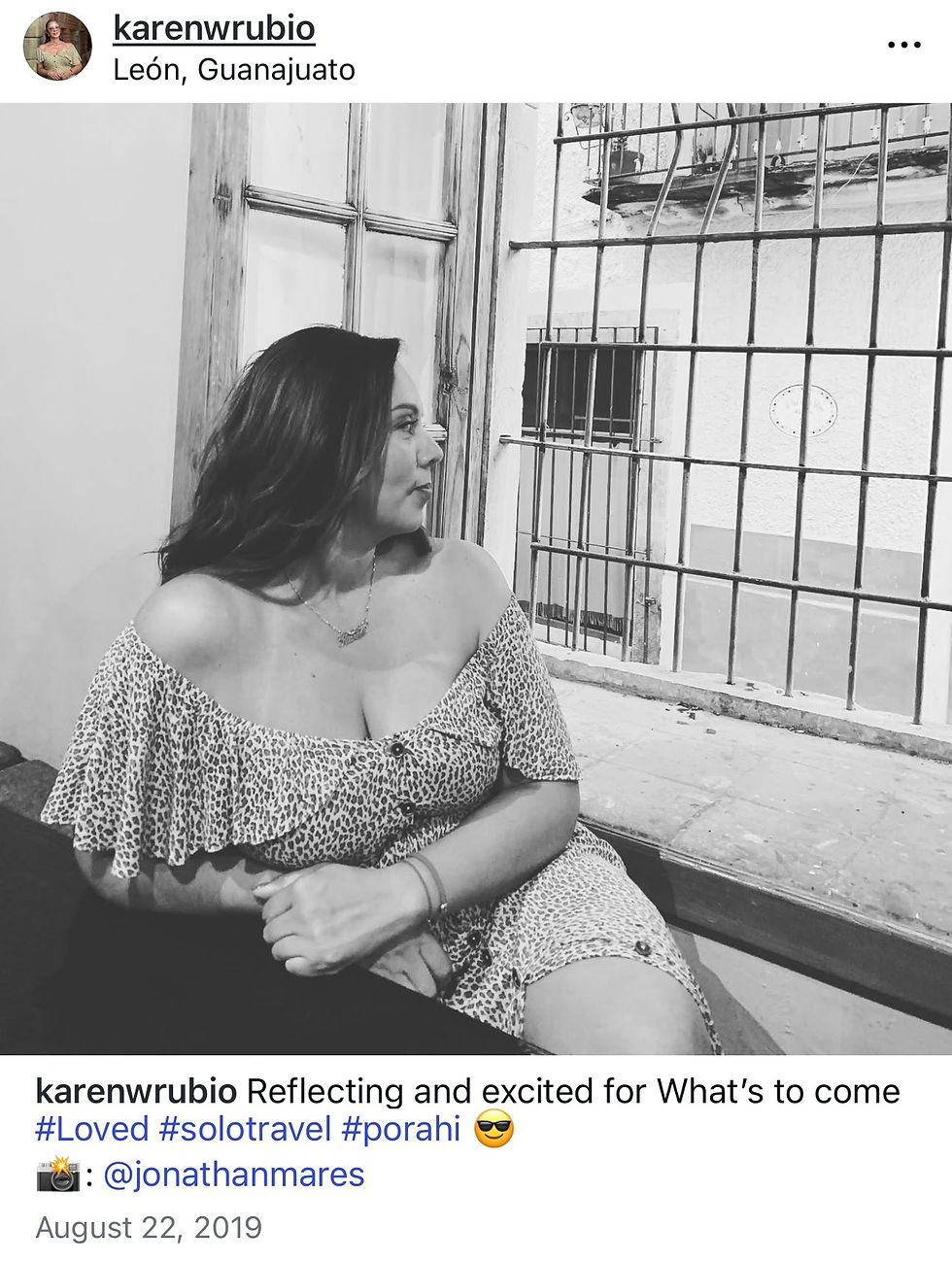 Black-and-white photo of a woman in a leopard-print dress sitting indoors by a barred window, looking away. Her body posture is guarded, with arms crossed in front, expressing a sense of heaviness and reflection.