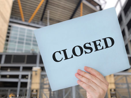 Hand holding a sign that reads ‘CLOSED’ in front of a modern hospital building, symbolizing rural hospital closures.
