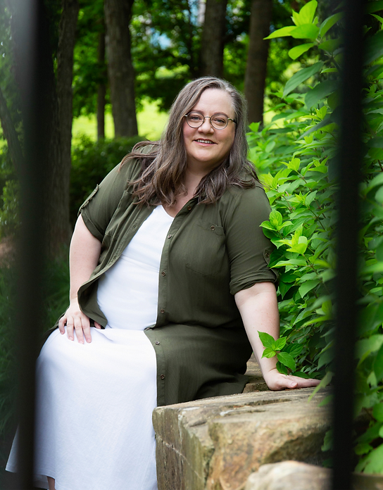 This is an image of me sitting on a stone bench surrounded by greenery. I am smiling & my hand is on the stone bench, as if to invite you to come talk to me.