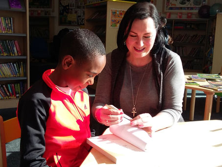 Angelina Kalahari and a boy sit at a table in a library. She points at a book, smiling. Shelves of colorful books are visible in the well-lit room.