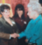 Three women in a formal setting, one in a turquoise outfit shaking hands with another in a dark suit. Smiles and warm expressions.