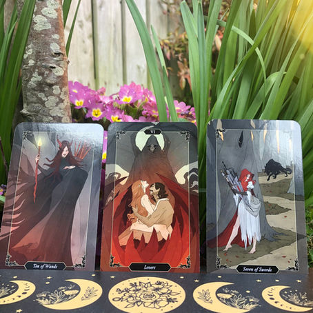 Three tarot cards on display in a garden setting: Lovers, Ten of Wands, Seven of Swords. Vibrant flowers and greenery surround them.