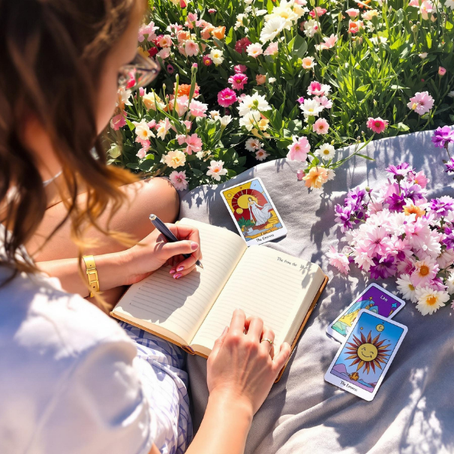 A woman writes in a notebook on a blanket, surrounded by tarot cards and colorful flowers. Sunny outdoor setting, peaceful mood.