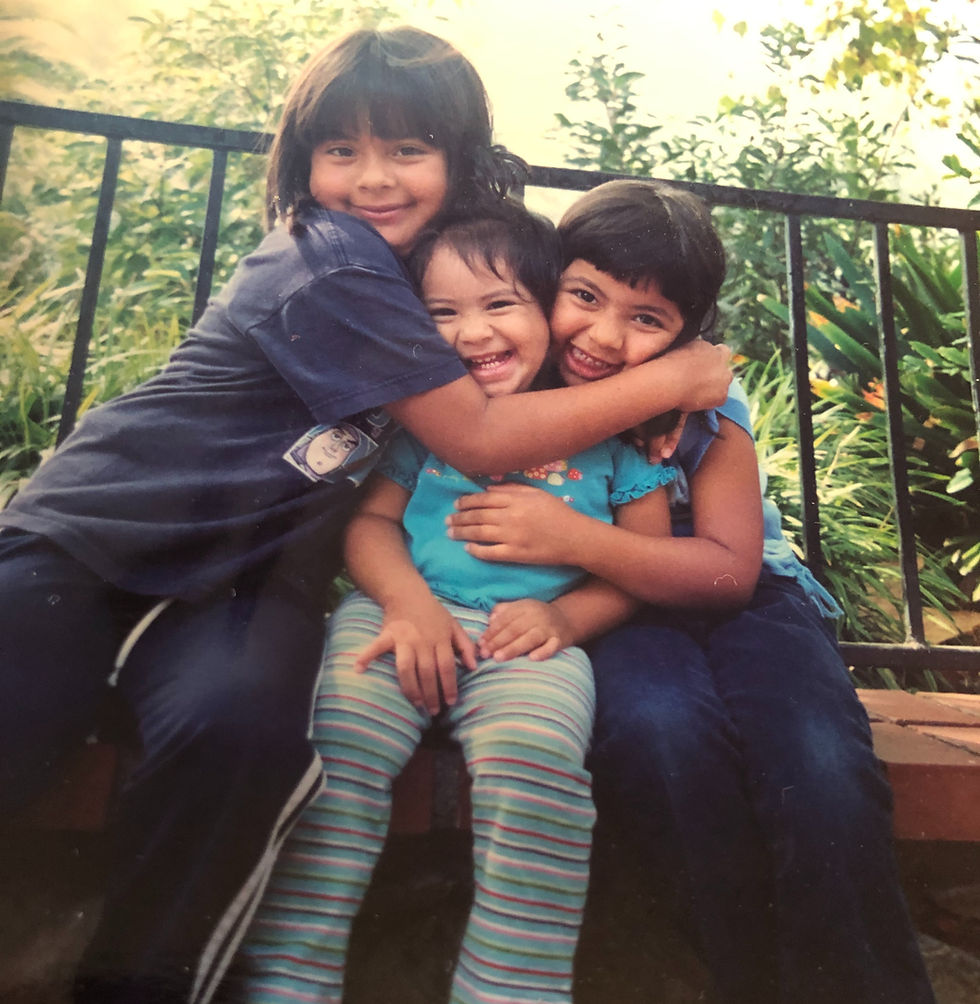 Three children sitting on a bench, smiling and hugging each other. They're wearing colorful clothes. Greenery and a fence are in the background.