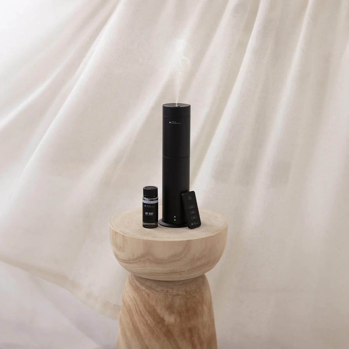Black Hotel Collection smart scent diffuser releasing mist, styled on a sculptural wooden pedestal beside fragrance oil bottles, evoking a luxury hotel ambiance