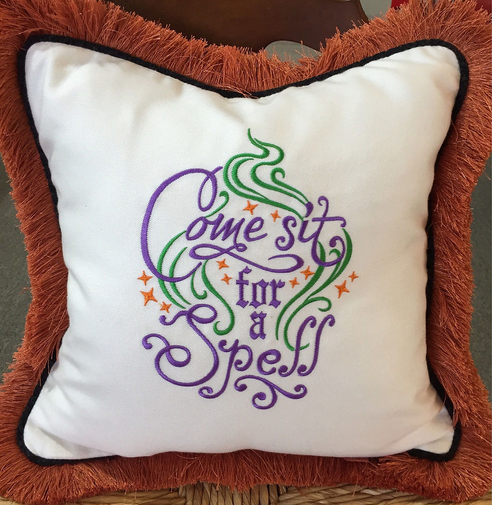 "Come sit for a spell" embroidered on white pillow with orange brush fringe around edges.
